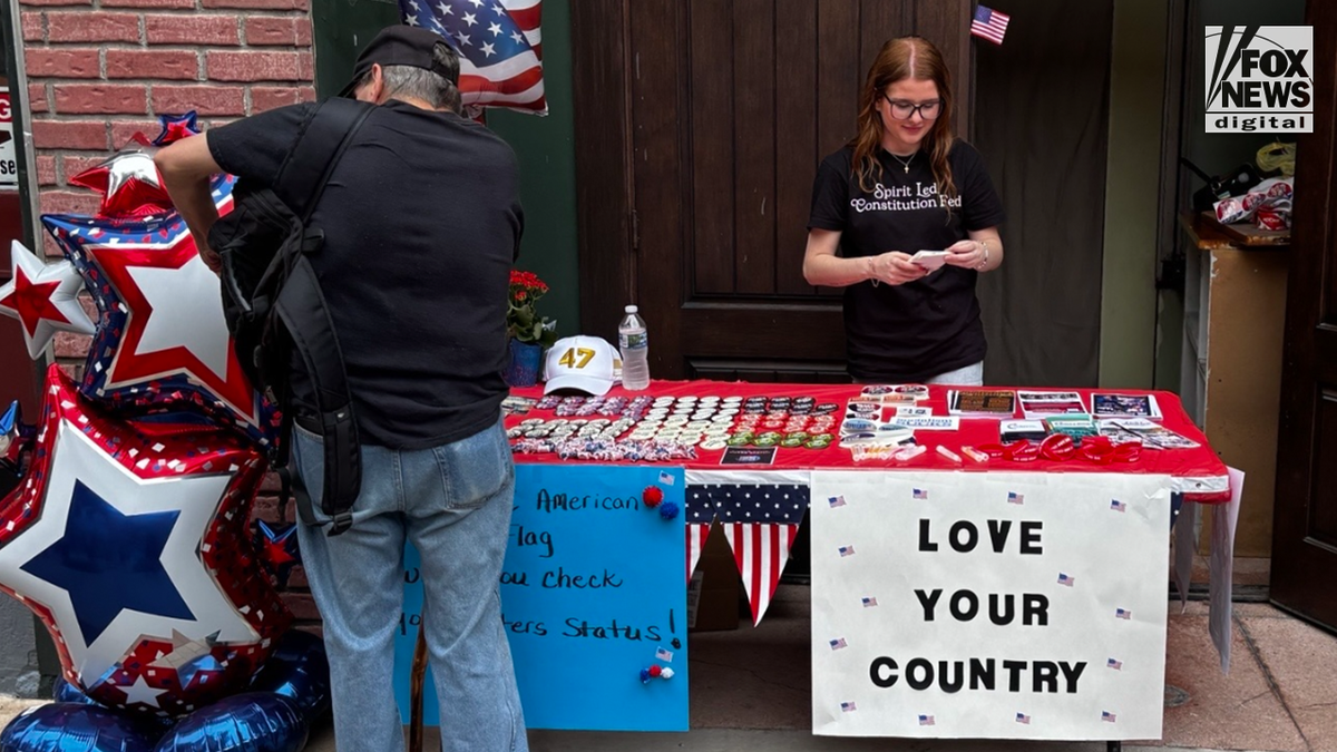 A volunteer stands behind a display table with buttons, flags and a "Love Your Country" sign as a visitor browses during the America 250 celebration in Cumberland, Maryland.