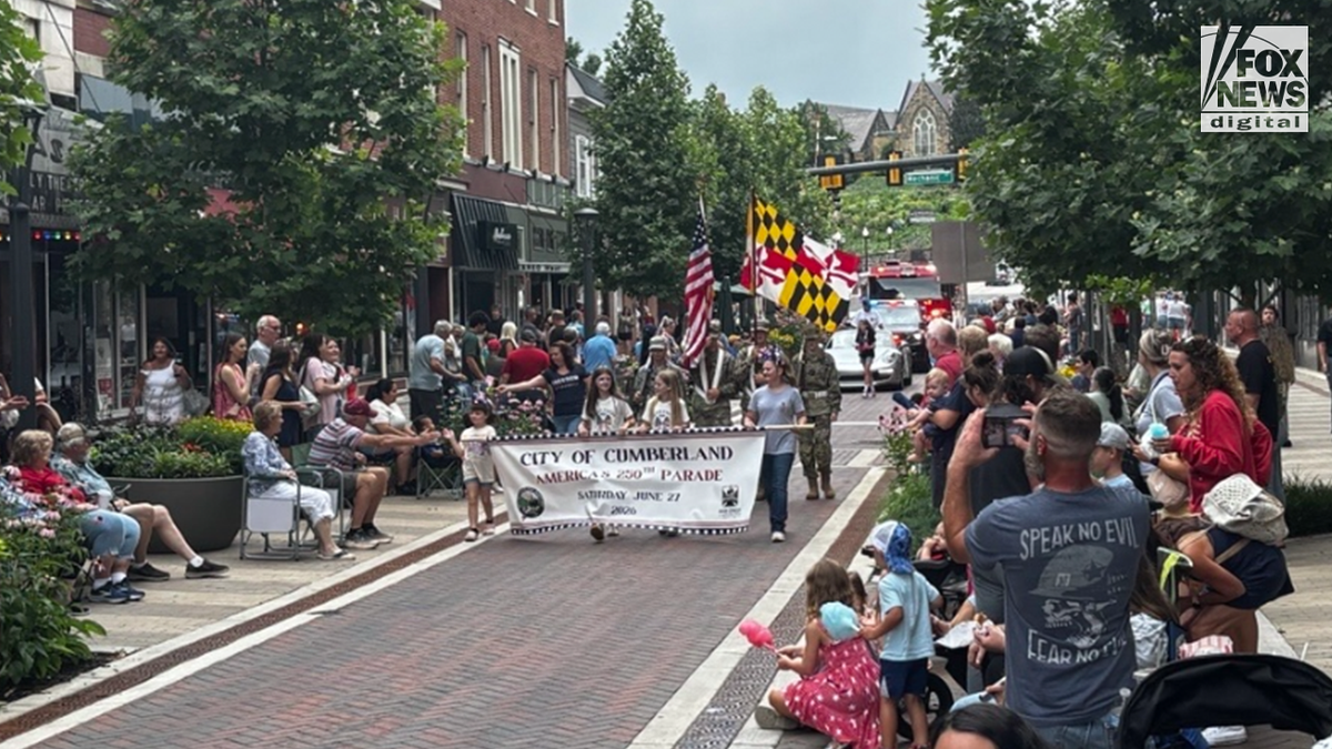Parade participants carry the City of Cumberland America 250 banner and American and Maryland flags in downtown Cumberland, Maryland, as spectators line both sides of the street.