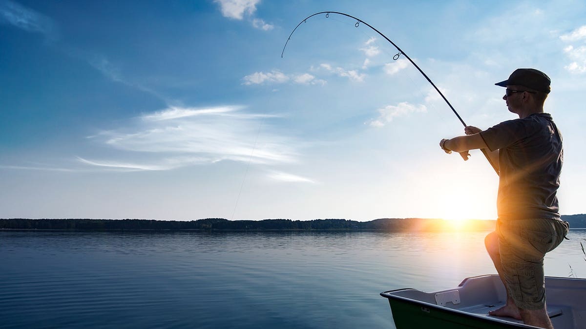 A fisherman holding a fishing rod standing by a lake at sunset.