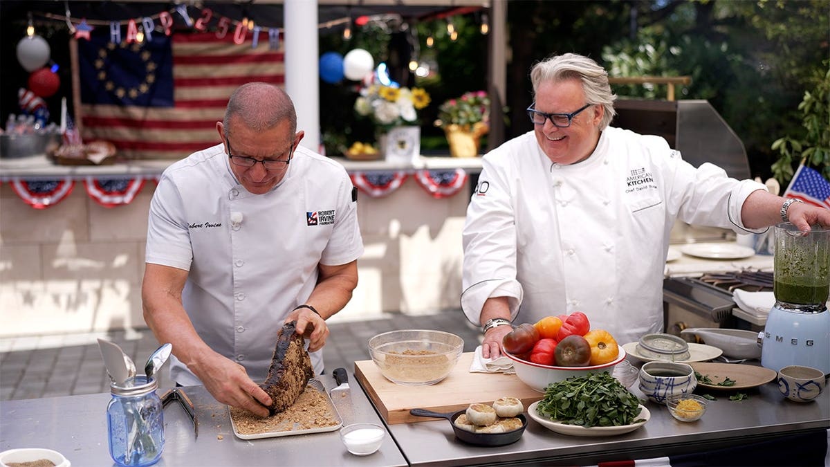 Chef Robert Irvine and Chef David Burke prepare a steak together during an outdoor cooking demonstration.