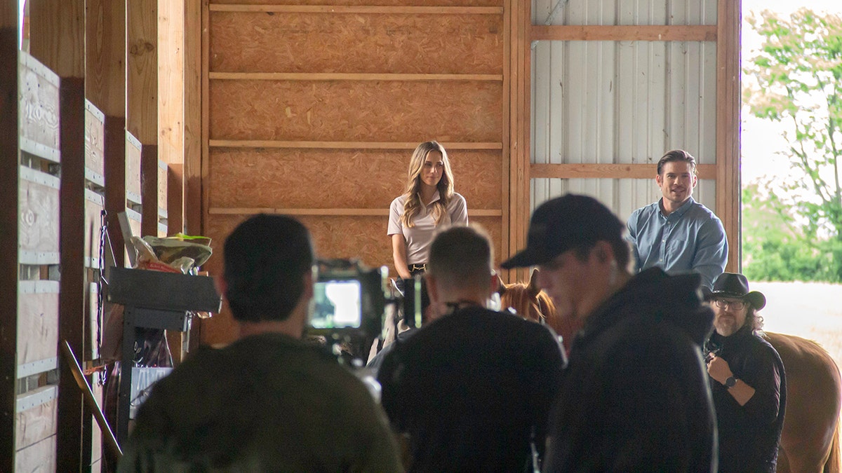 Film crew working inside a horse barn while two riders sit on horseback near an open doorway.