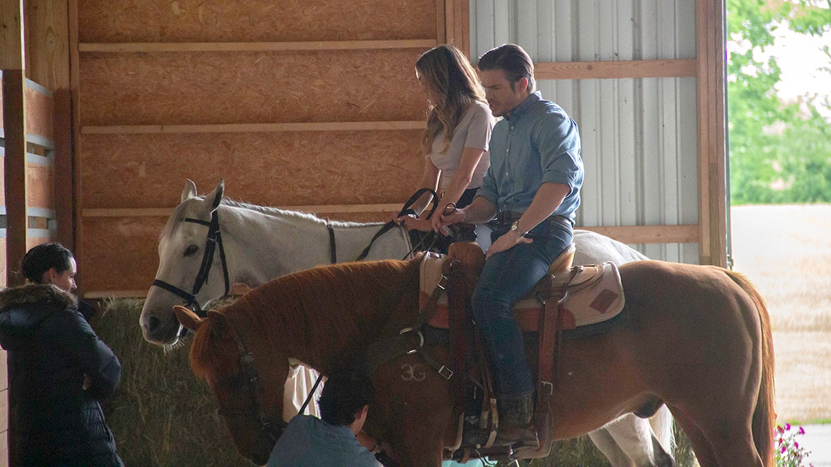 Jana Kramer and co-star seated on horses inside a barn as crew members prepare equipment nearby.