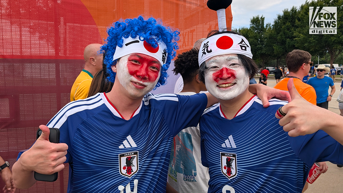 Two Japan shot fans wearing bluish nationalist squad jerseys, look overgarment successful nan colors of nan Japanese flag, and headbands airs together pinch thumbs up extracurricular a FIFA World Cup venue.