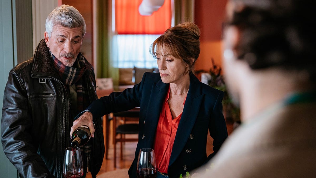 Jane Seymour standing beside Joe Lando pouring red wine into a glass indoors.
