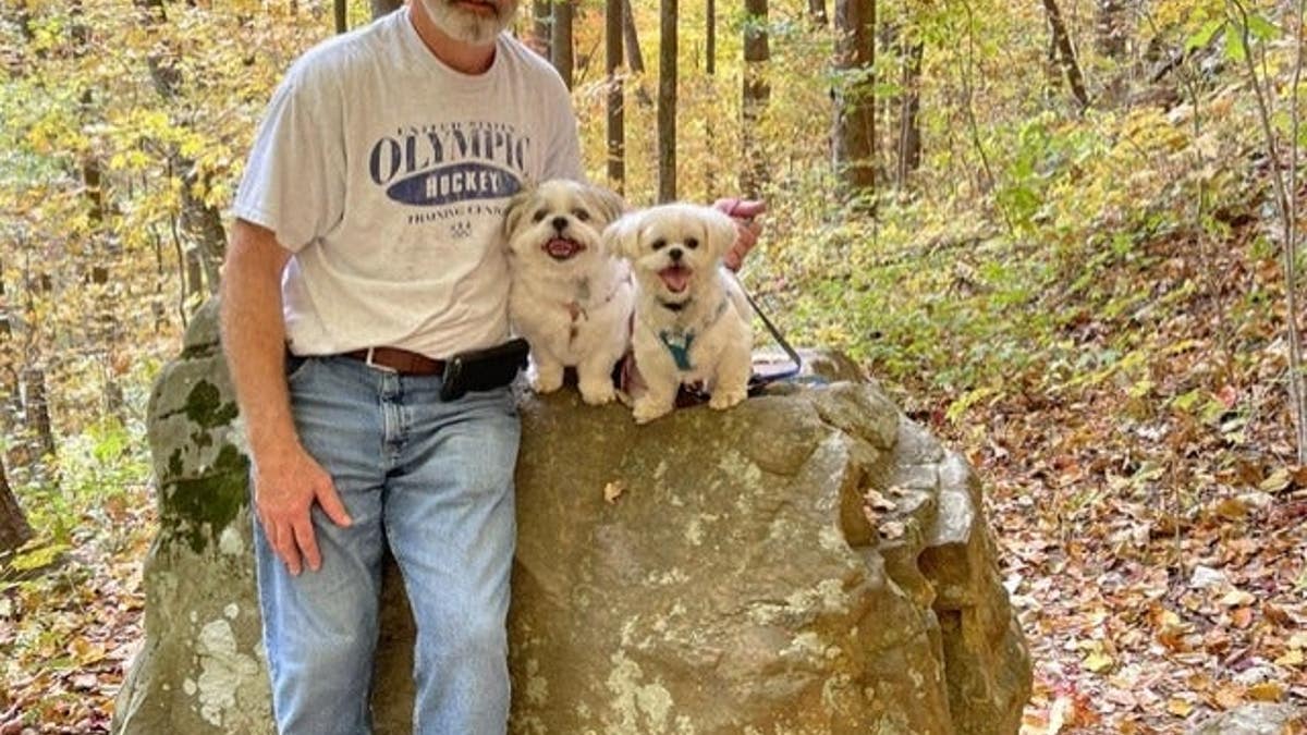 man in T-shirt and jeans stands next to a rock in the woods with two small white dogs