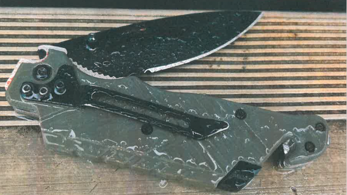 A folding knife with a dark blade and gray handle rests on a wooden surface, shown in a close-up image with water droplets visible across the knife.