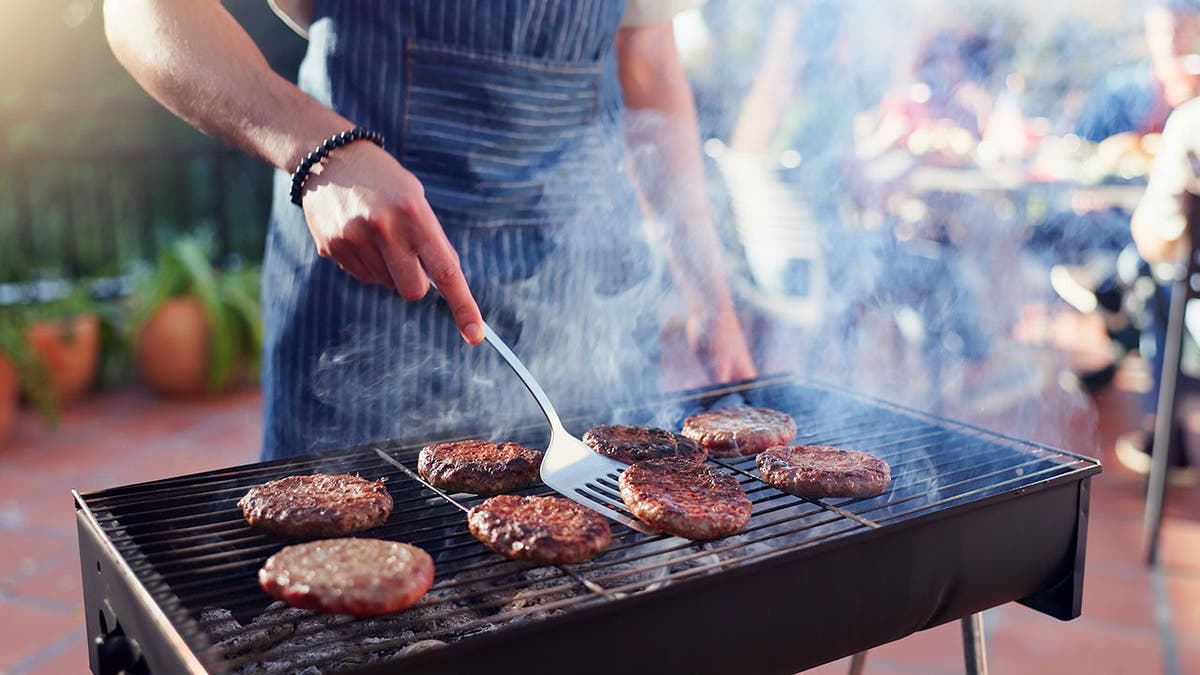 A man waring an apron grills hamburgers.