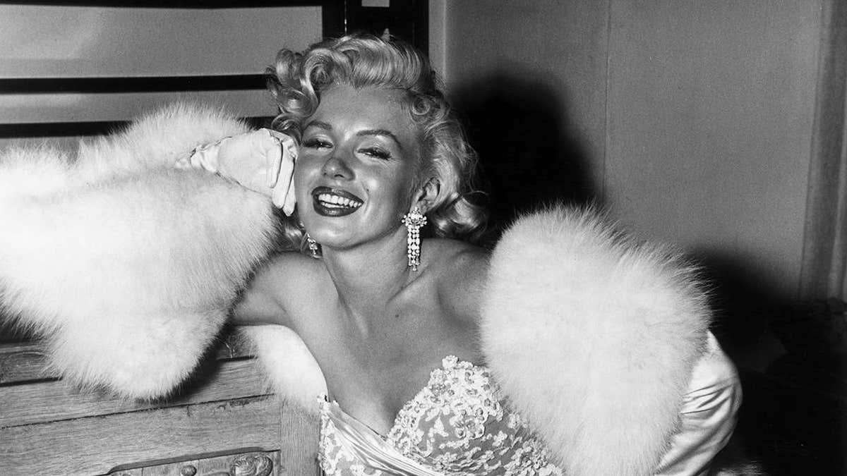 Marilyn Monroe smiling and sitting wearing a lace evening gown and white fur stole.