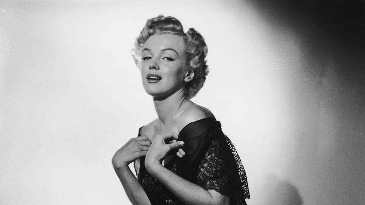 Marilyn Monroe kneeling on an animal fur wearing a black negligee in a studio portrait.