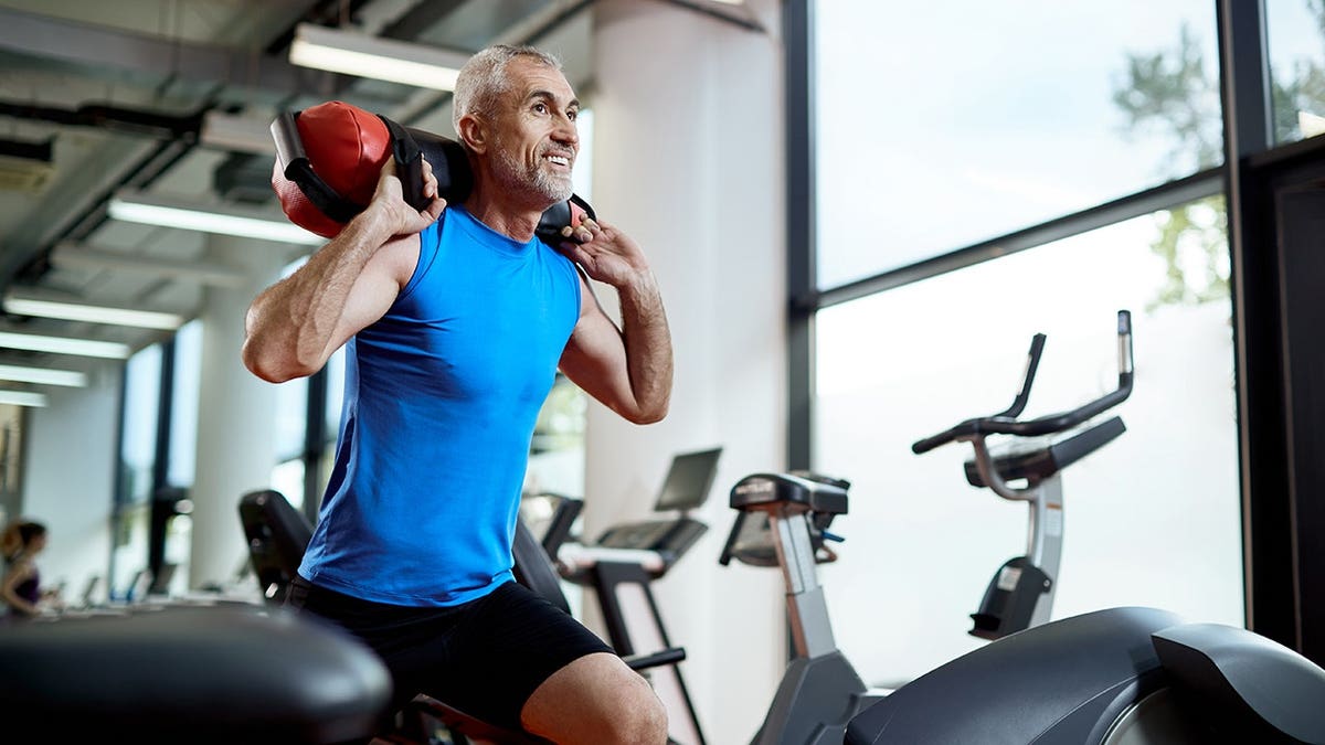 Happy middle aged athlete carrying sandbag on his shoulders while working out in a gym