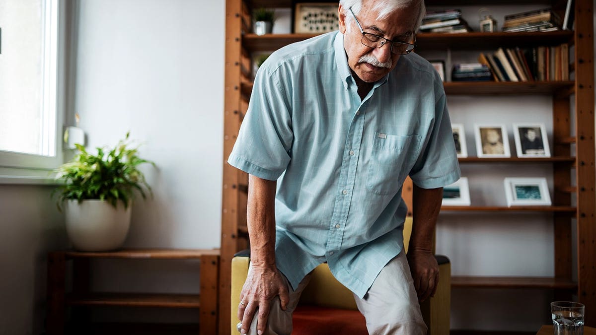 Senior adult man holding his knee while standing in a living room.