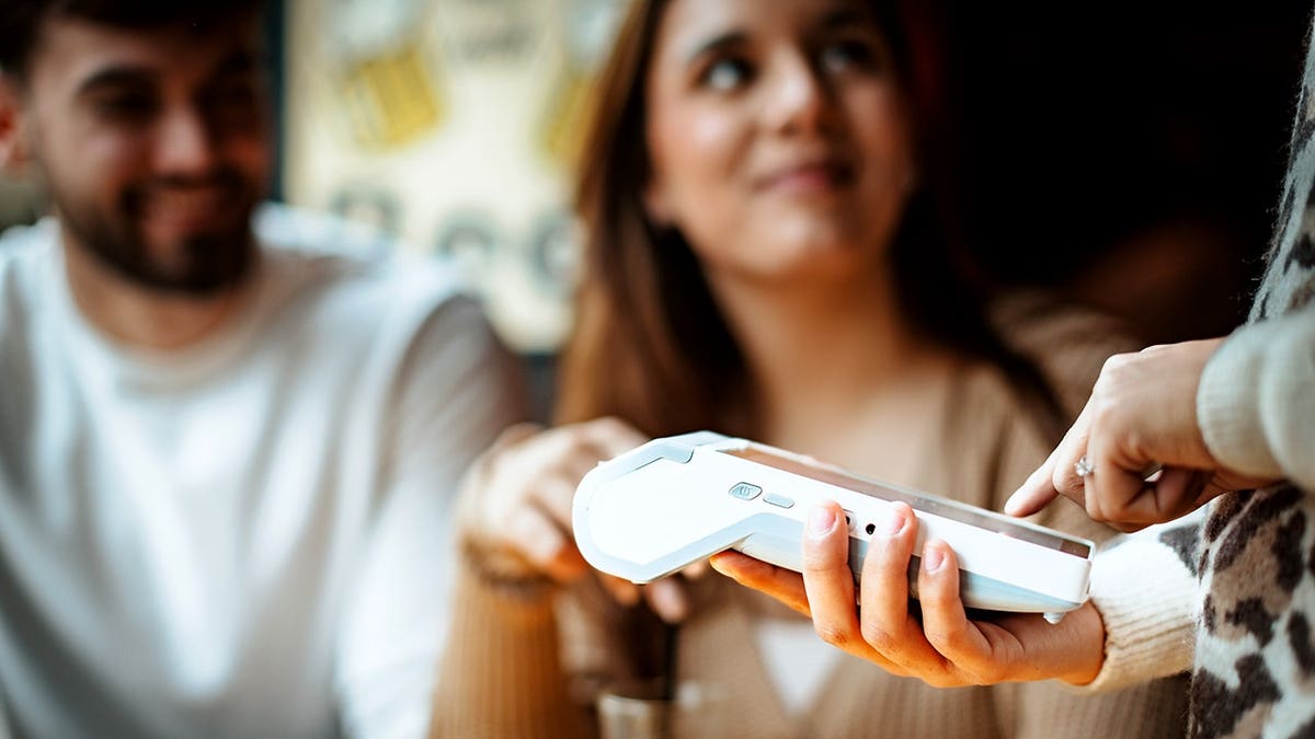 A server handing a payment device to a customer at a cafe with two friends sitting nearby.