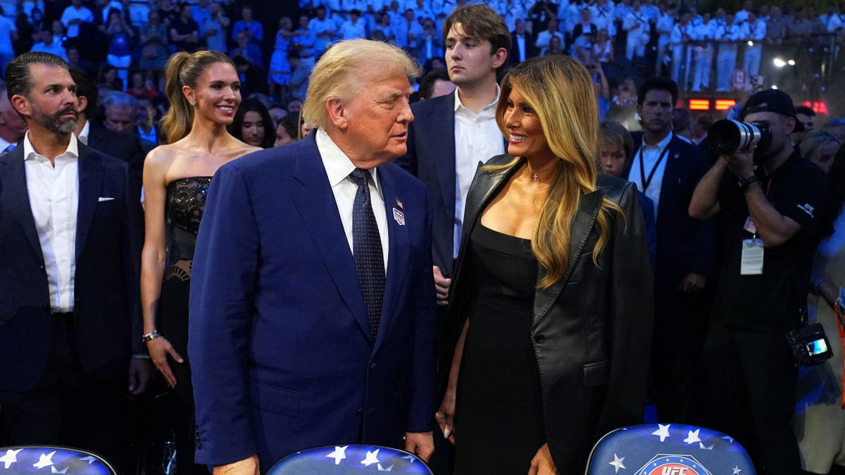 O presidente Donald J. Trump e a primeira-dama dos Estados Unidos Melania Trump sorriem no evento UFC Freedom 250. Donald Trump Jr., sua esposa Bettina Anderson e Barron Trump estão atrás deles.