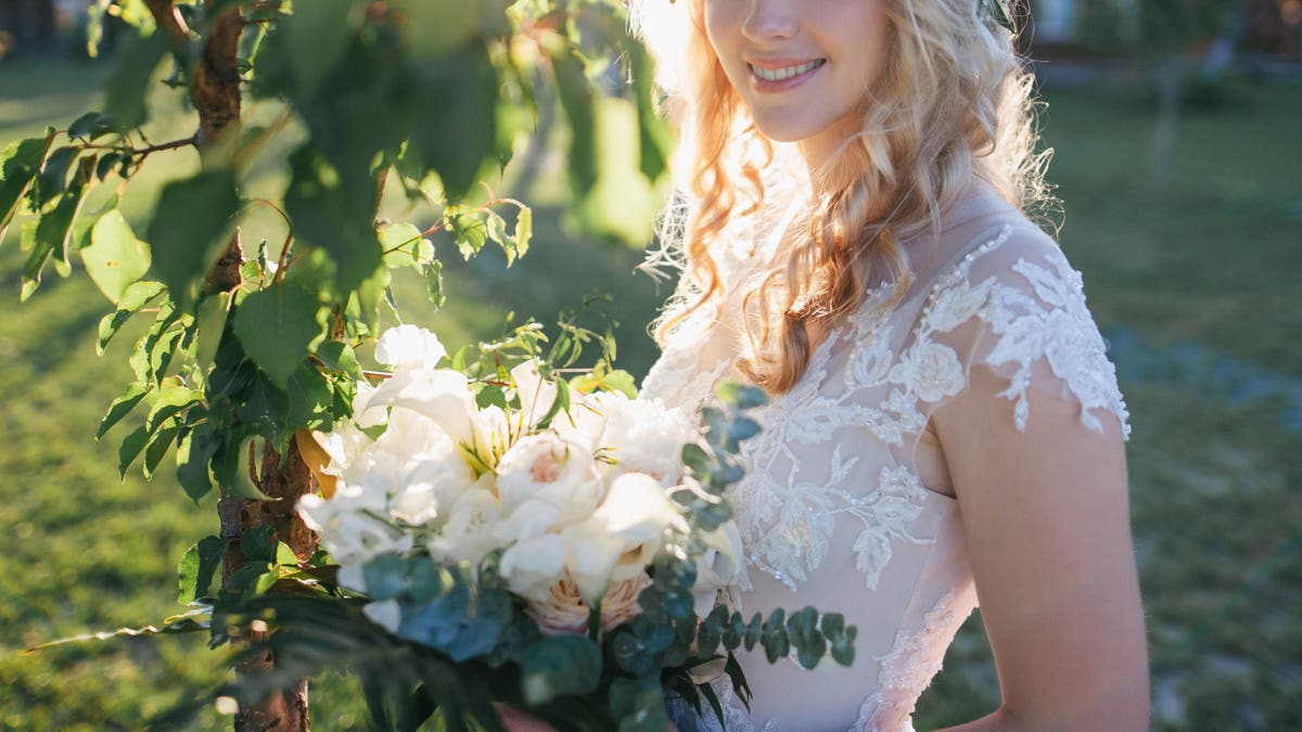 Young bride in wedding dress and floral wreath holding bouquet and smiling at camera