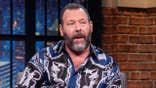 Comedian Bert Kreischer sitting and talking with host Seth Meyers on a late night show set