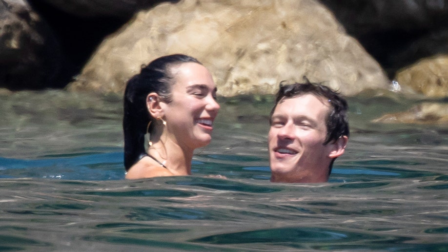 Dua Lipa and Callum Turner smiling while swimming together in Italy.