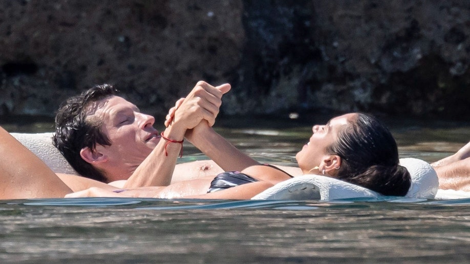 Dua Lipa and Callum Turner holding hands while floating in the ocean.