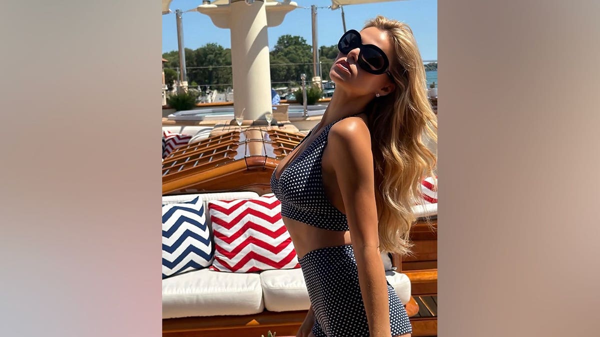 Brooks Nader tilts her head back while posing on the deck of a yacht in Venice wearing a navy-and-white polka dot matching set.