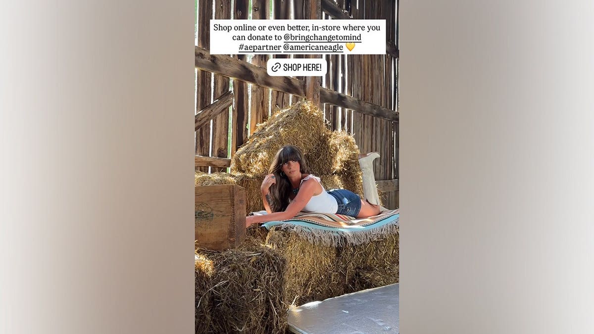Ella Langley lies across stacked hay bales while wearing a white tank top, dark-wash denim shorts and white cowboy boots during an American Eagle photoshoot.