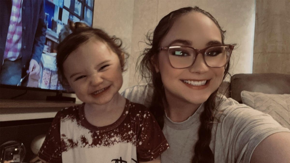 Woman with glasses and a toddler girl smile together for a selfie in a living room.