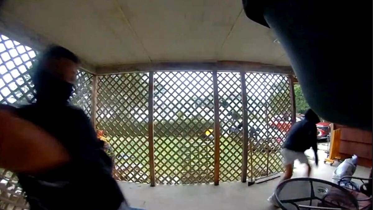 Fisheye porch-camera still showing two people in dark clothing and face coverings on a lattice-enclosed porch, one at left and one at right appearing to move away, with a yard and parked vehicles visible through the lattice.