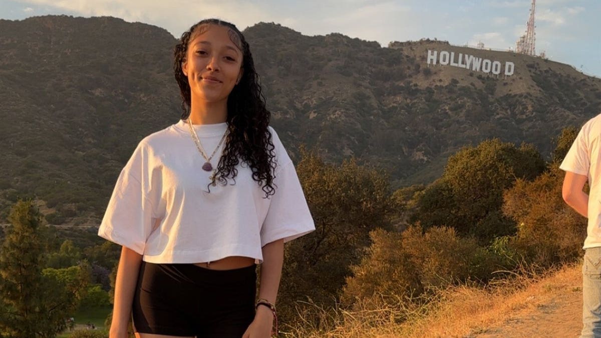 Yoseliani Ann Marte Ocasio, a young woman in a white T-shirt, stands smiling outdoors with the Hollywood sign on a hillside behind her.