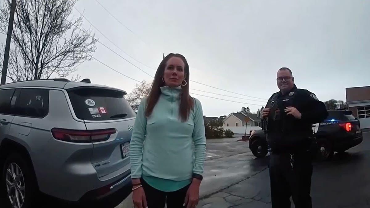 A woman in a yellow jacket is standing in the parking lot next to a police officer and patrol cars with their lights on.