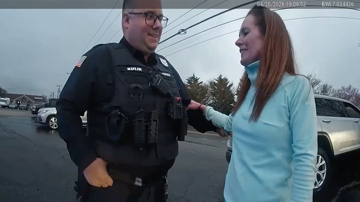 A body camera still shows a police officer standing with a woman in a yellow jacket in a parking lot, the time stamp is visible on the screen.