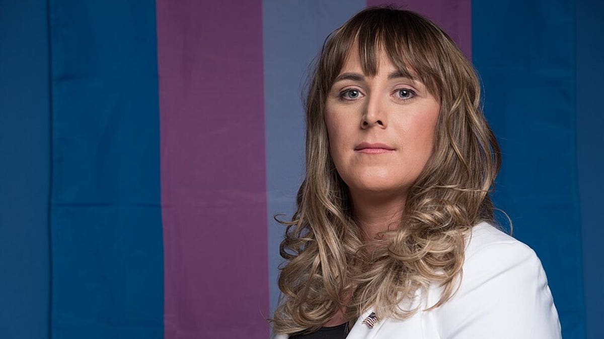 Woman with long wavy brown hair wearing a white blazer stands in front of a blue, pink and white transgender pride flag.