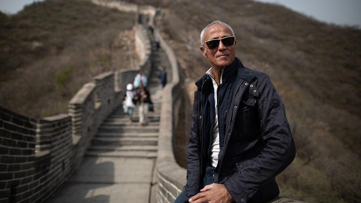 Andrew Ridgeley sits on the Great Wall of China looking into the distance with visitors walking behind him.