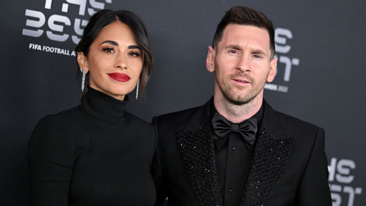 Antonella Roccuzzo and Lionel Messi pose together on a green carpet.