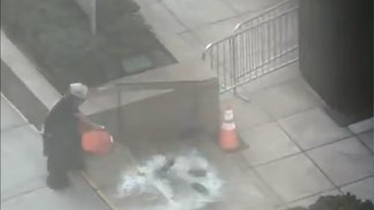 A man in black with a white helmet dumps an orange bucket of gasoline onto the steps outside the building