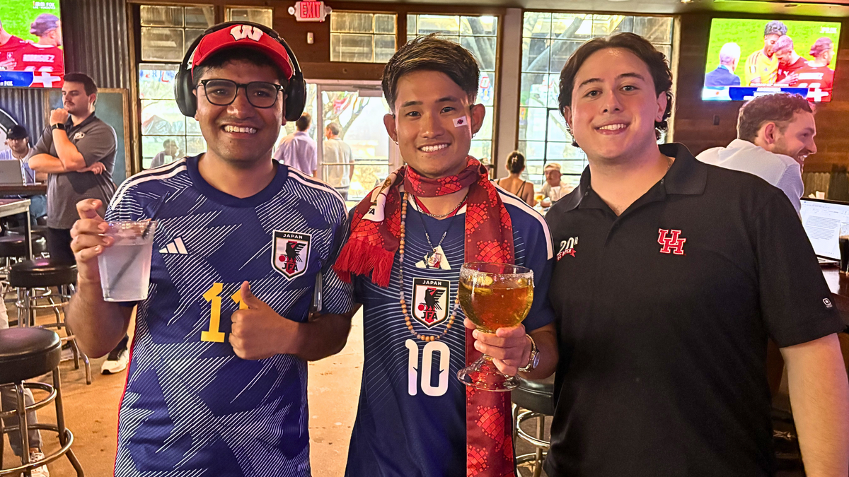 Un influencer japonés de las redes sociales posa en un Dallas , con aficionados al fútbol a su izquierda y a su derecha, sonriendo mientras sostiene unas copas y posa para la cámara.