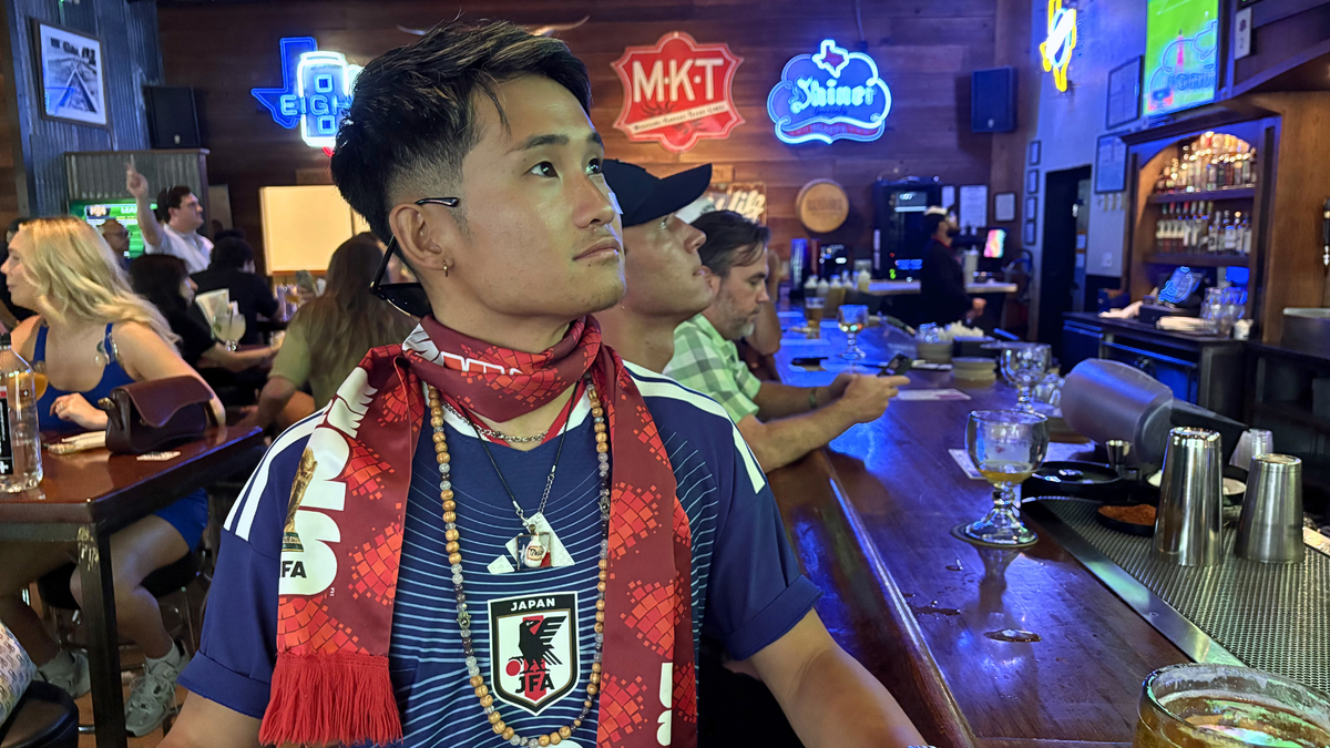 Un hombre asiático mira hacia otro lado, hacia la tele que tiene enfrente, mientras está sentado en un bar de Texas letreros de neón al fondo