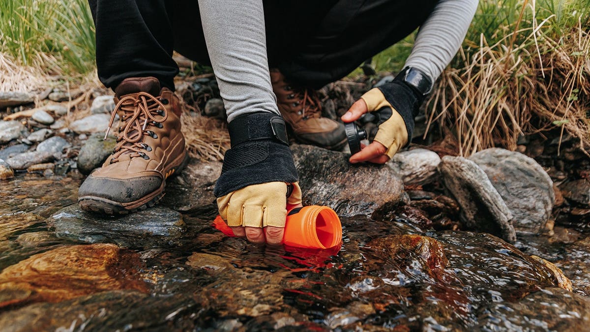 Hiker kneels beside a flowing stream and fills a water bottle in the woods.