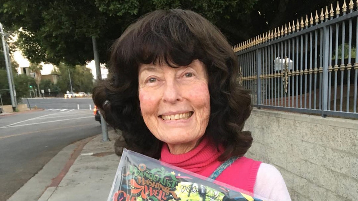 Linda Perhacs smiles while holding a vinyl record in a clear sleeve on a quiet sidewalk.