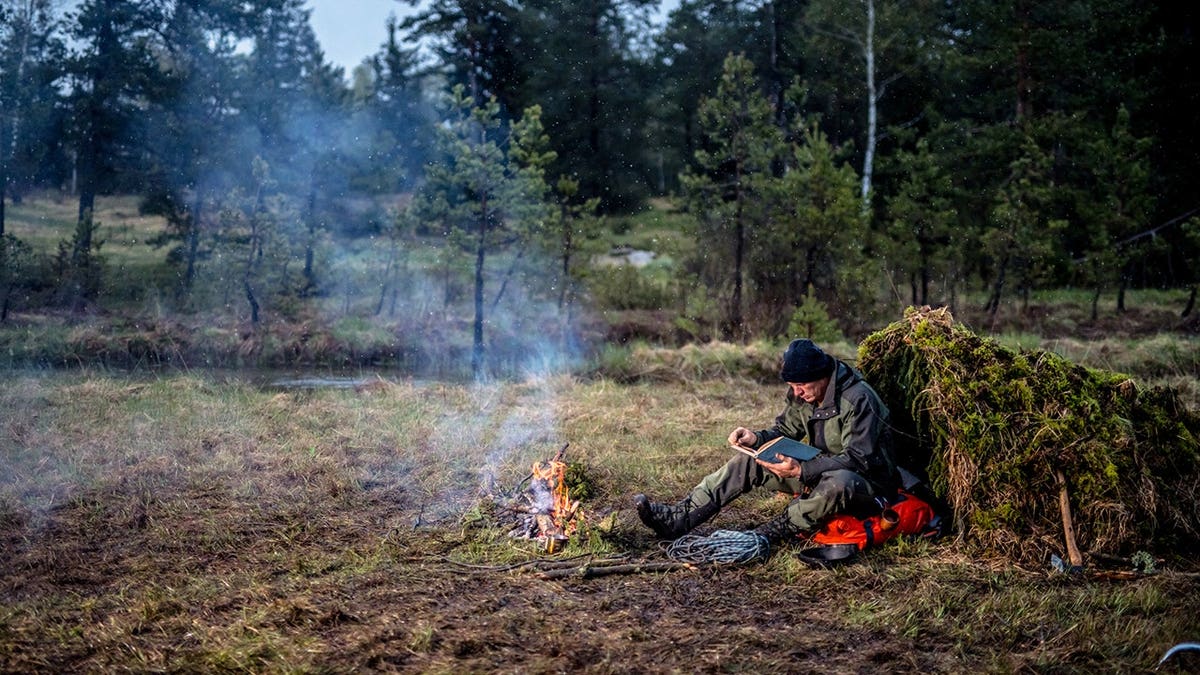 Man reads a book while sitting near a campfire in a forest.
