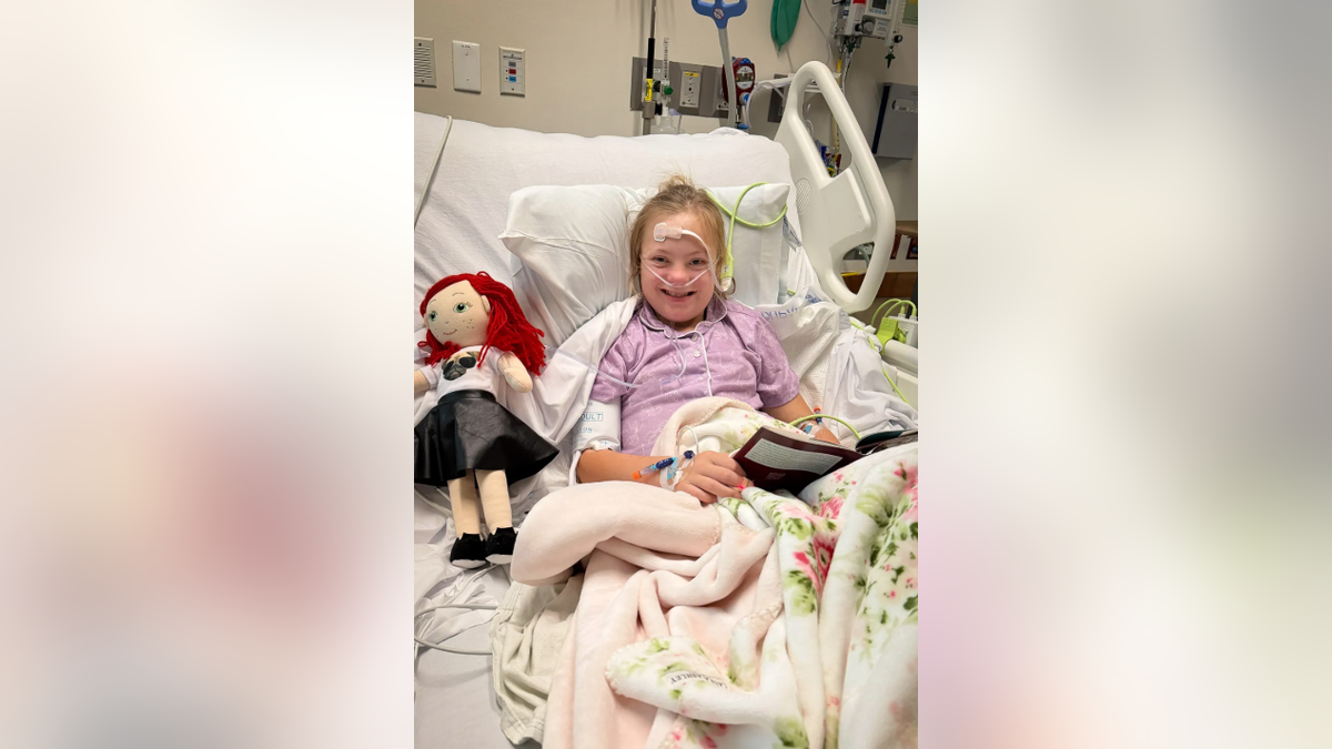 Rory Feek's daughter smiles while lying in a hospital bed holding a book.