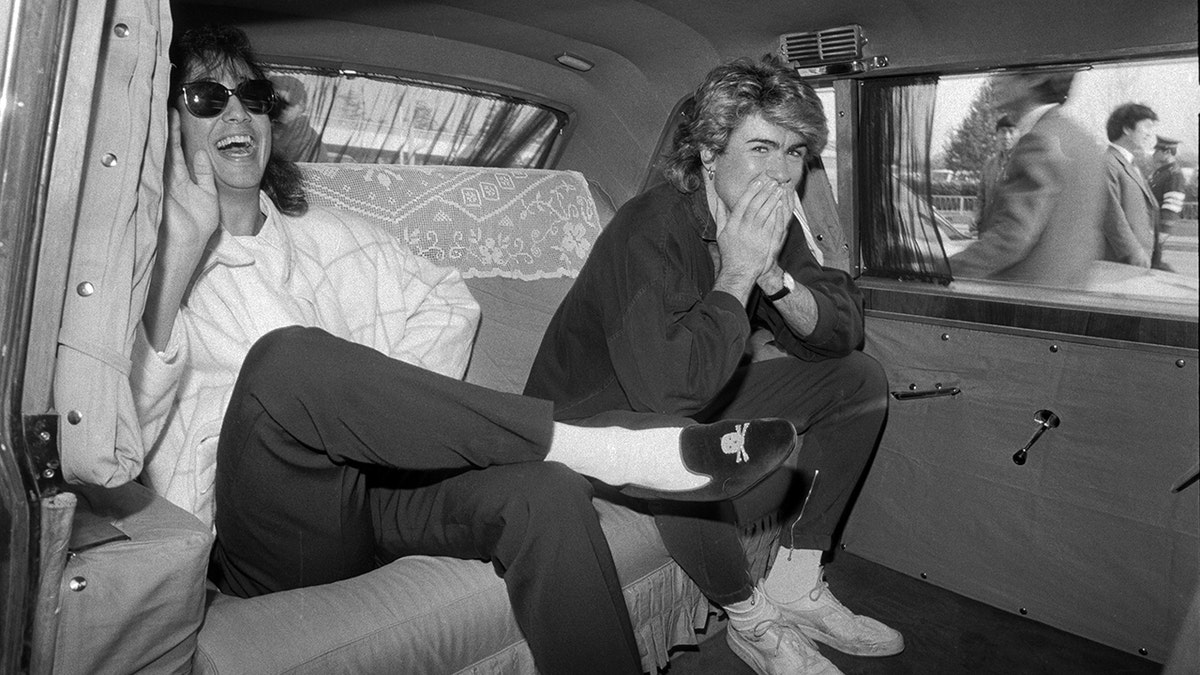 George Michael and Andrew Ridgeley laugh and wave while seated inside a vehicle with lace-covered seats.