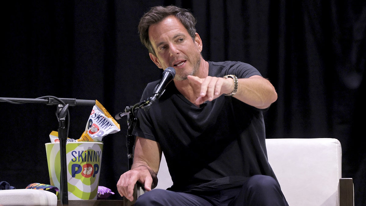 Will Arnett is pointing while seated next to a bucket of Skinny Pop.