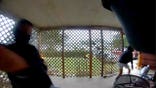 Fisheye porch-camera still showing two people in dark clothing and face coverings on a lattice-enclosed porch, one at left and one at right appearing to move away, with a yard and parked vehicles visible through the lattice.