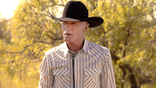 Ed Harris stands outdoors wearing a black cowboy hat and striped western shirt.