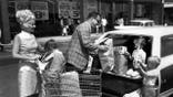 Man loads groceries into a station wagon as two boys help and a girl sits in a shopping cart.