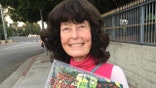 Linda Perhacs smiles while holding a vinyl record in a clear sleeve on a quiet sidewalk.