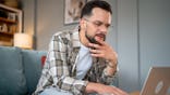 Man with beard and glasses types on laptop while sitting on sofa.