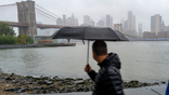 A man walks through Brooklyn during heavy rain and high winds with the NYC skyline in the background.