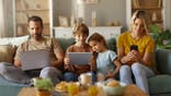 Parents and children use wireless devices while sitting on a sofa in a living room.