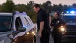 Two police officers stand with a driver beside a car on a scenic road at sunset.