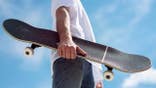 Young man holds a skateboard outdoors against a blue sky with clouds.