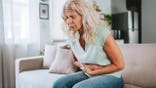 Woman sits on a sofa holding her stomach with a painful expression.